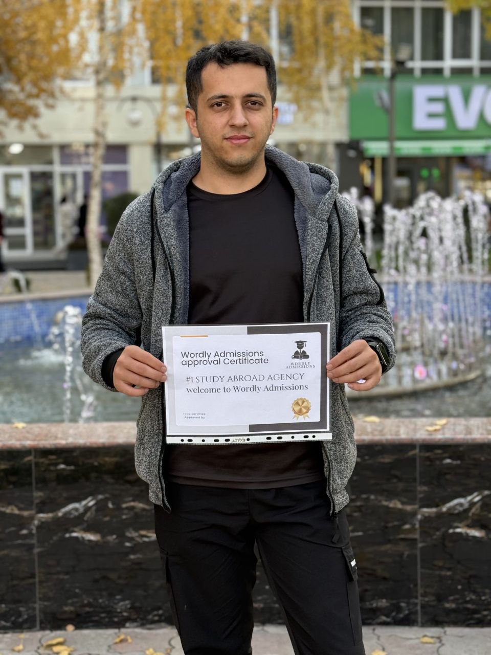 Wordly Admissions student holding his approval certificate by a city fountain